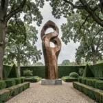 Sculpture géante en bronze abstrait trônant dans un jardin paysager avec haies de buis et allée de gravier
