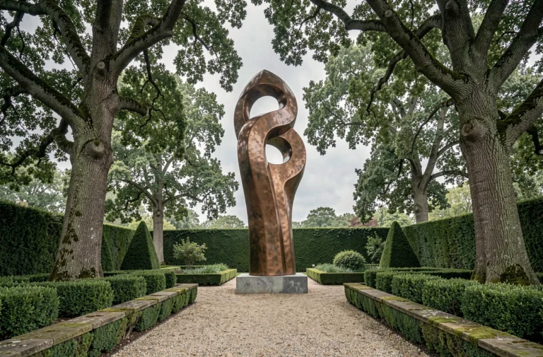Sculpture géante en bronze abstrait trônant dans un jardin paysager avec haies de buis et allée de gravier